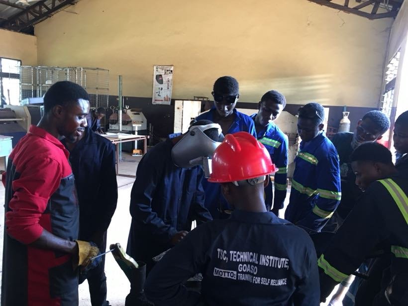 Students in overalls gathered around an instructor in the workshop